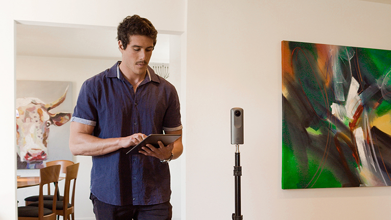 Man standing behind tripod with a 360 degree camera holding an iPad in a living room